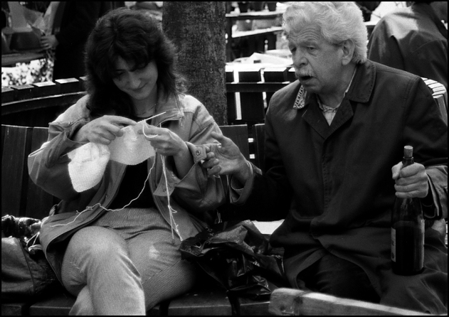 junge Frau mit Strickzeug, alter Mann mit Rotweinflasche im Gespräch