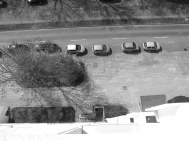 Schwarz-Weißbild. Blick aus dem neunten Stock nach unten: Fußweg, geparkte Autos, Bäume, Straße, Schatten